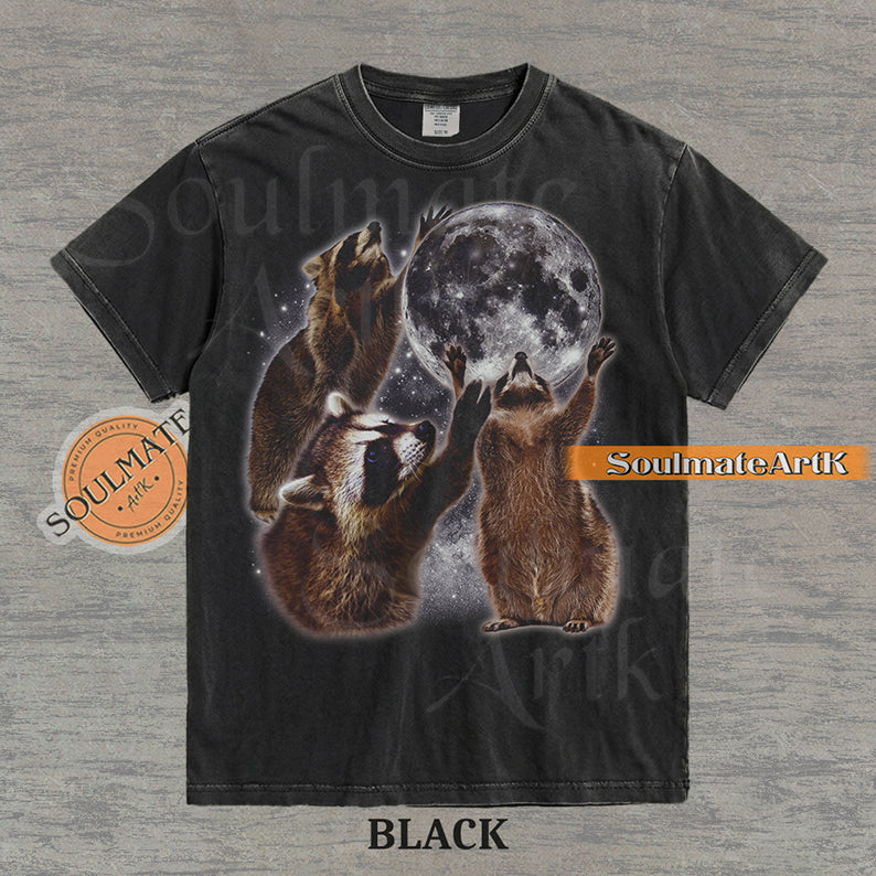 Graphic featuring three detailed raccoons reaching for a giant full moon in a starry space setting on a washed black t-shirt.