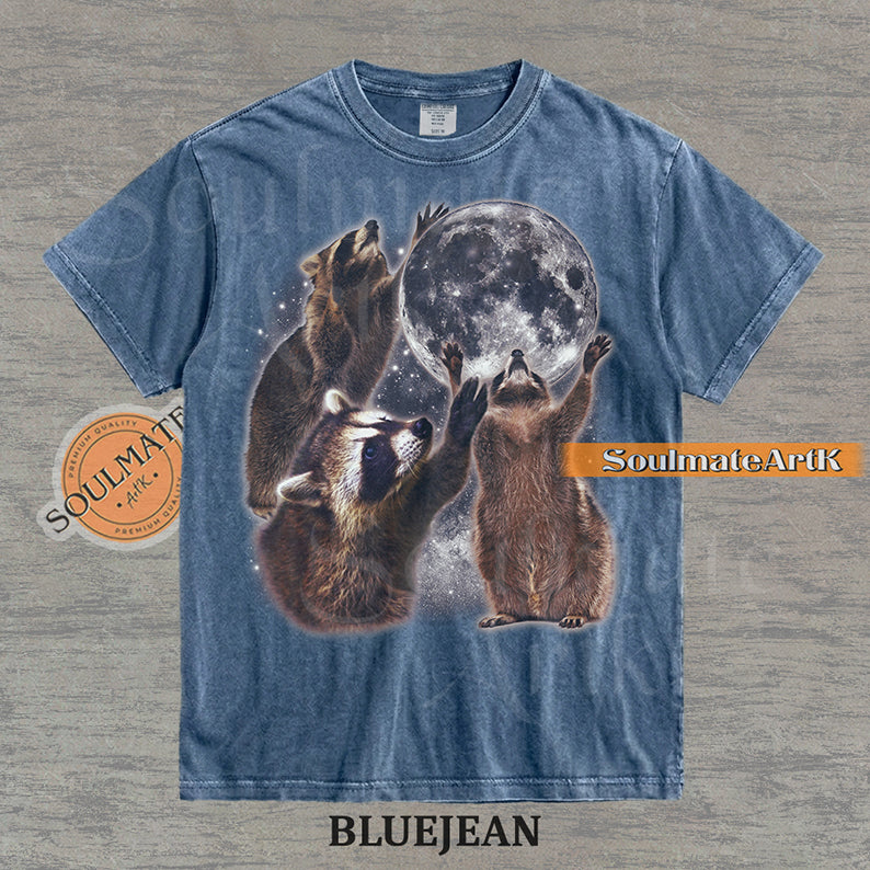 Graphic featuring three detailed raccoons reaching for a giant full moon in a starry space setting on a washed bluejean t-shirt.