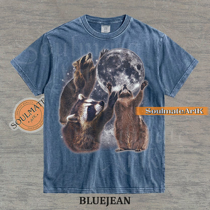 Graphic featuring three detailed raccoons reaching for a giant full moon in a starry space setting on a washed bluejean t-shirt.