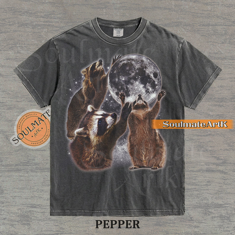 Graphic featuring three detailed raccoons reaching for a giant full moon in a starry space setting on a washed pepper t-shirt.