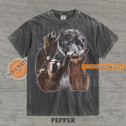 Graphic featuring three detailed raccoons reaching for a giant full moon in a starry space setting on a washed pepper t-shirt.