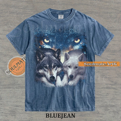 Vintage-style celestial graphic of a timber wolf and arctic wolf resting beneath large glowing wolf eyes in a starry sky on a bluejean t-shirt.
