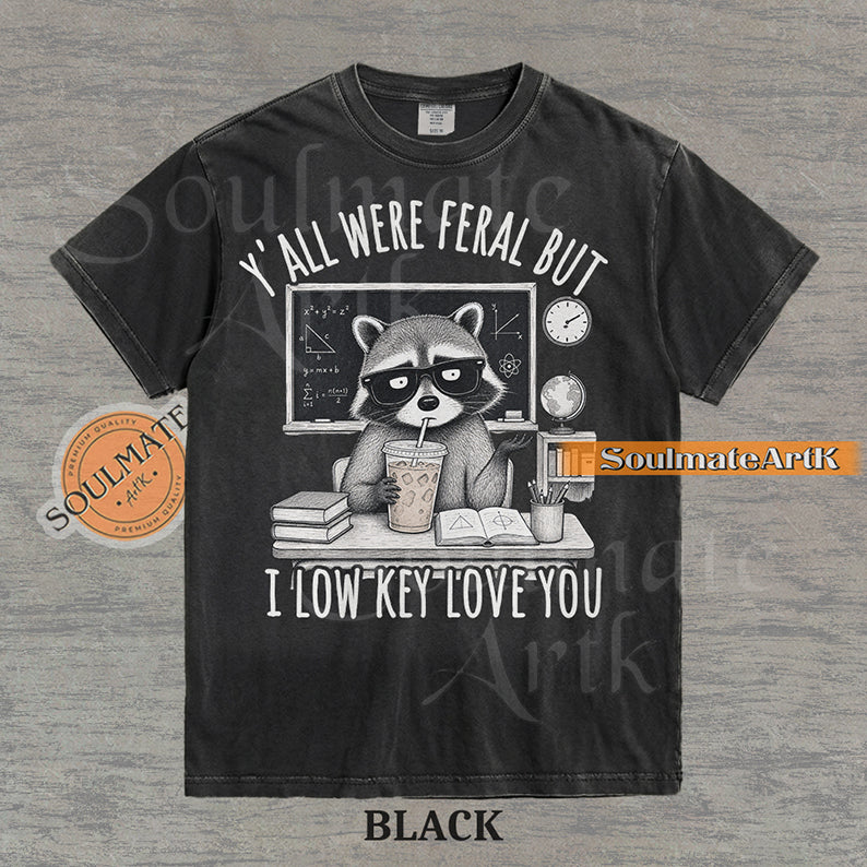 Hand-drawn illustration of a raccoon teacher at a desk with coffee and books under text reading "Y'all Were Feral But I Low Key Love You" on a washed black shirt.