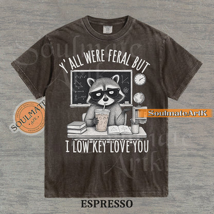 Hand-drawn illustration of a raccoon teacher at a desk with coffee and books under text reading "Y'all Were Feral But I Low Key Love You" on a washed espresso shirt.