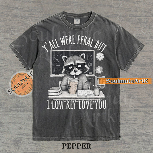 Hand-drawn illustration of a raccoon teacher at a desk with coffee and books under text reading "Y'all Were Feral But I Low Key Love You" on a washed pepper shirt.
