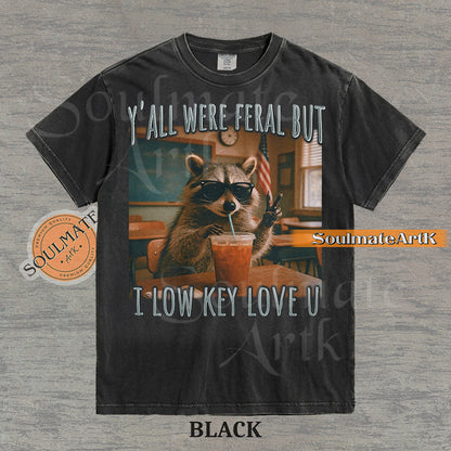Photorealistic graphic of a raccoon teacher in sunglasses with iced coffee in a classroom under text reading "Y'all Were Feral But I Low Key Love U" on a washed black shirt.