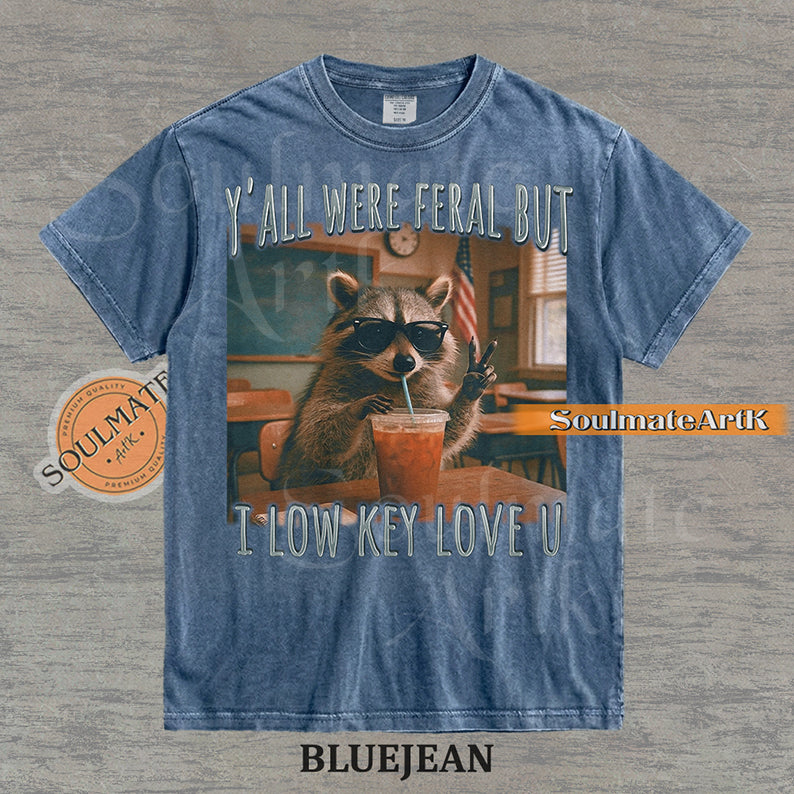 Photorealistic graphic of a raccoon teacher in sunglasses with iced coffee in a classroom under text reading "Y'all Were Feral But I Low Key Love U" on a washed bluejean shirt.