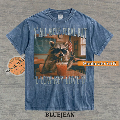 Photorealistic graphic of a raccoon teacher in sunglasses with iced coffee in a classroom under text reading "Y'all Were Feral But I Low Key Love U" on a washed bluejean shirt.