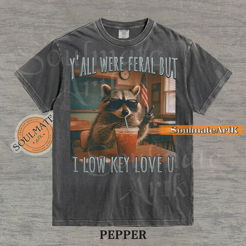 Photorealistic graphic of a raccoon teacher in sunglasses with iced coffee in a classroom under text reading "Y'all Were Feral But I Low Key Love U" on a washed pepper shirt.