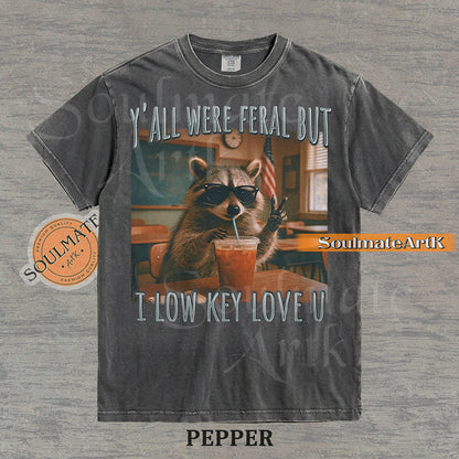 Photorealistic graphic of a raccoon teacher in sunglasses with iced coffee in a classroom under text reading "Y'all Were Feral But I Low Key Love U" on a washed pepper shirt.