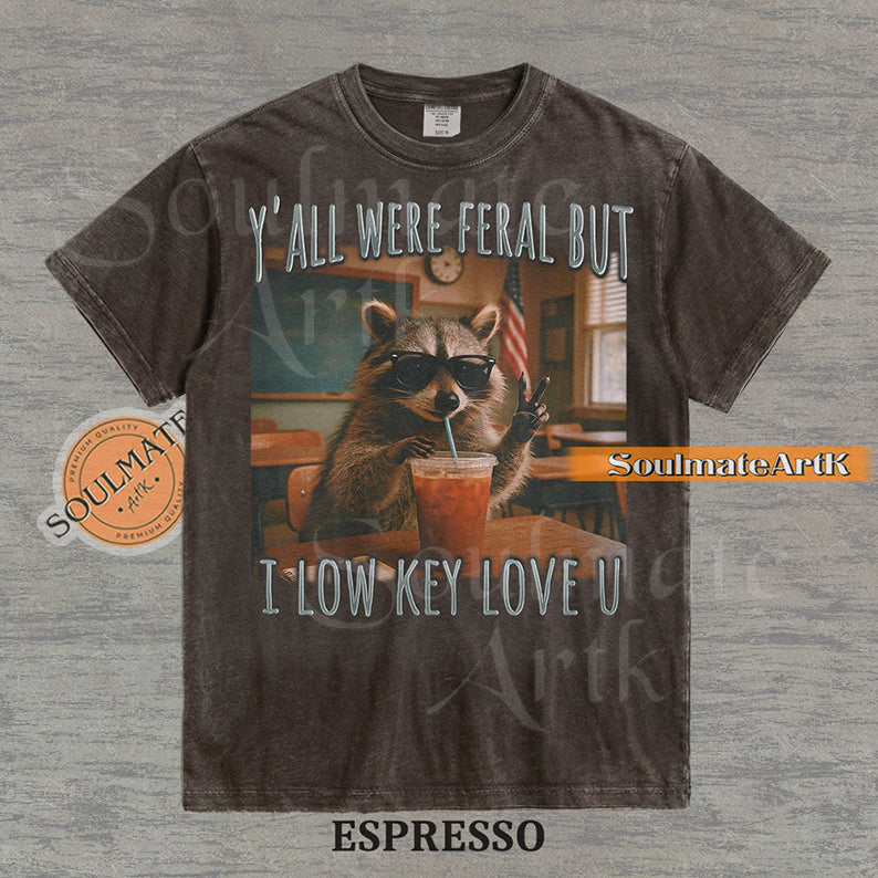 Photorealistic graphic of a raccoon teacher in sunglasses with iced coffee in a classroom under text reading "Y'all Were Feral But I Low Key Love U" on a washed espresso shirt.