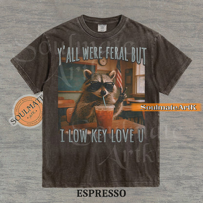 Photorealistic graphic of a raccoon teacher in sunglasses with iced coffee in a classroom under text reading "Y'all Were Feral But I Low Key Love U" on a washed espresso shirt.